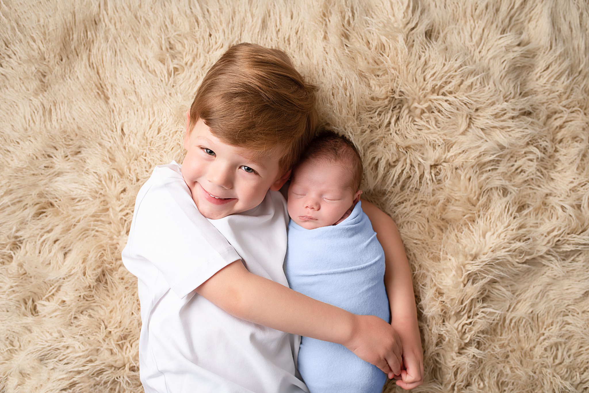 A newborn baby sleeps on a fur blanket in a blue swaddle while being hugged by his older brother