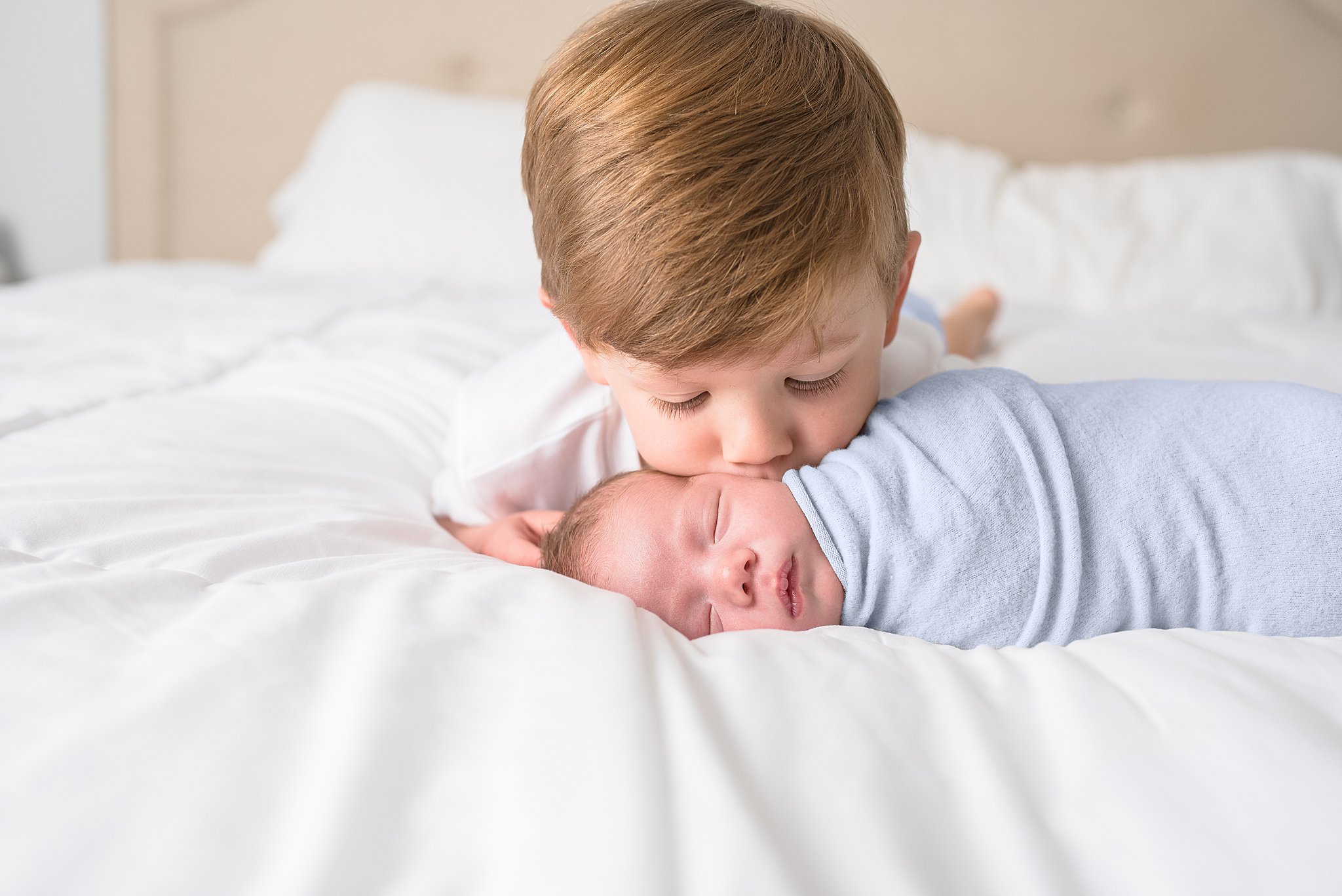 A newborn baby sleeps on a white bed with his older brother kissing his cheek San Diego Toy Stores