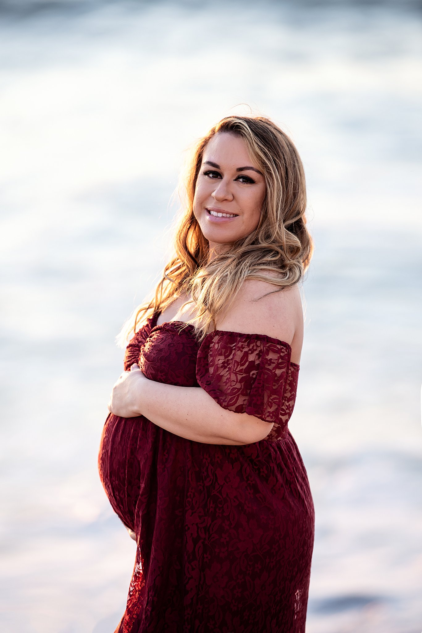 A mother to be holds her bump while wearing an off thee shoulder lace maternity gown on the beach