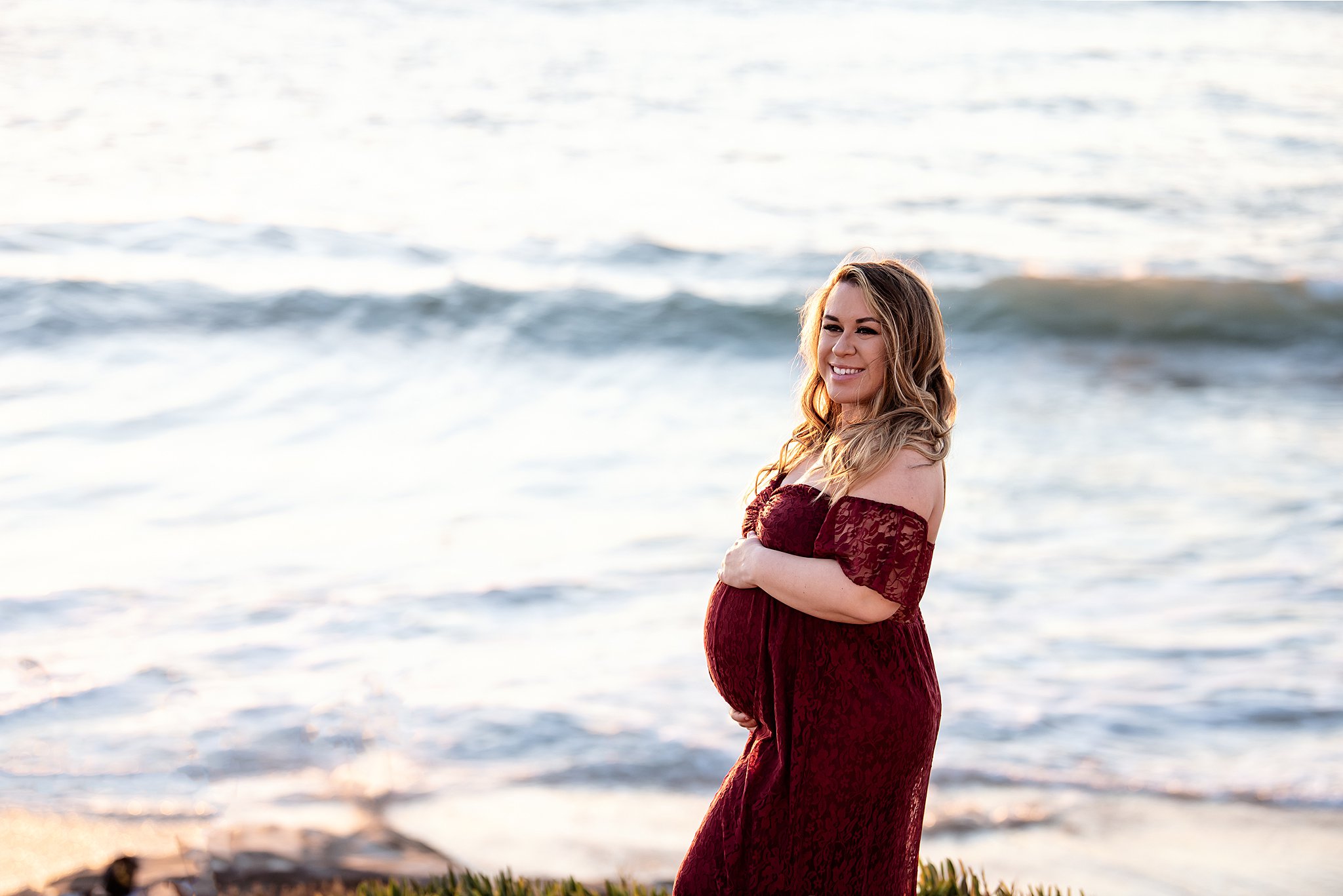 A mother to be in a dark red lace maternity dress stands on a beach while holding her bump Sharp Mary Birch Hospital for Women & Newborns