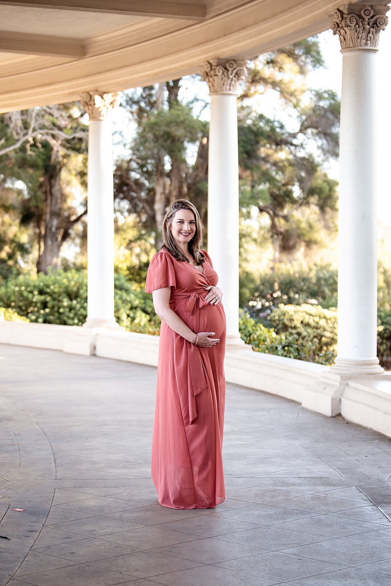 A mother to be stands amongst corinthian columns in a red maternity gown holding her bump