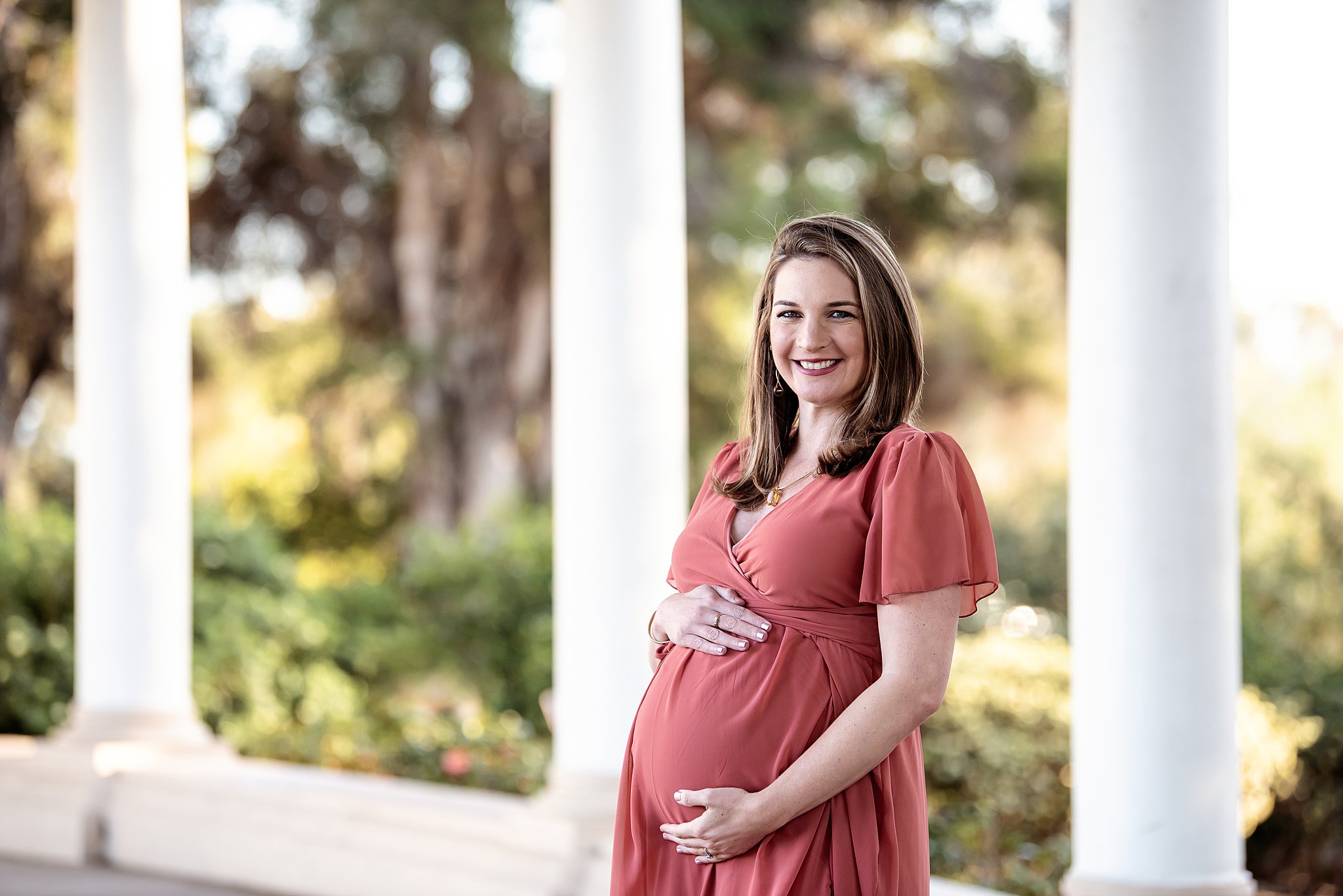 A mom to be stands in front of some pillars in a park while holding her bump Tourmaline Birth Center