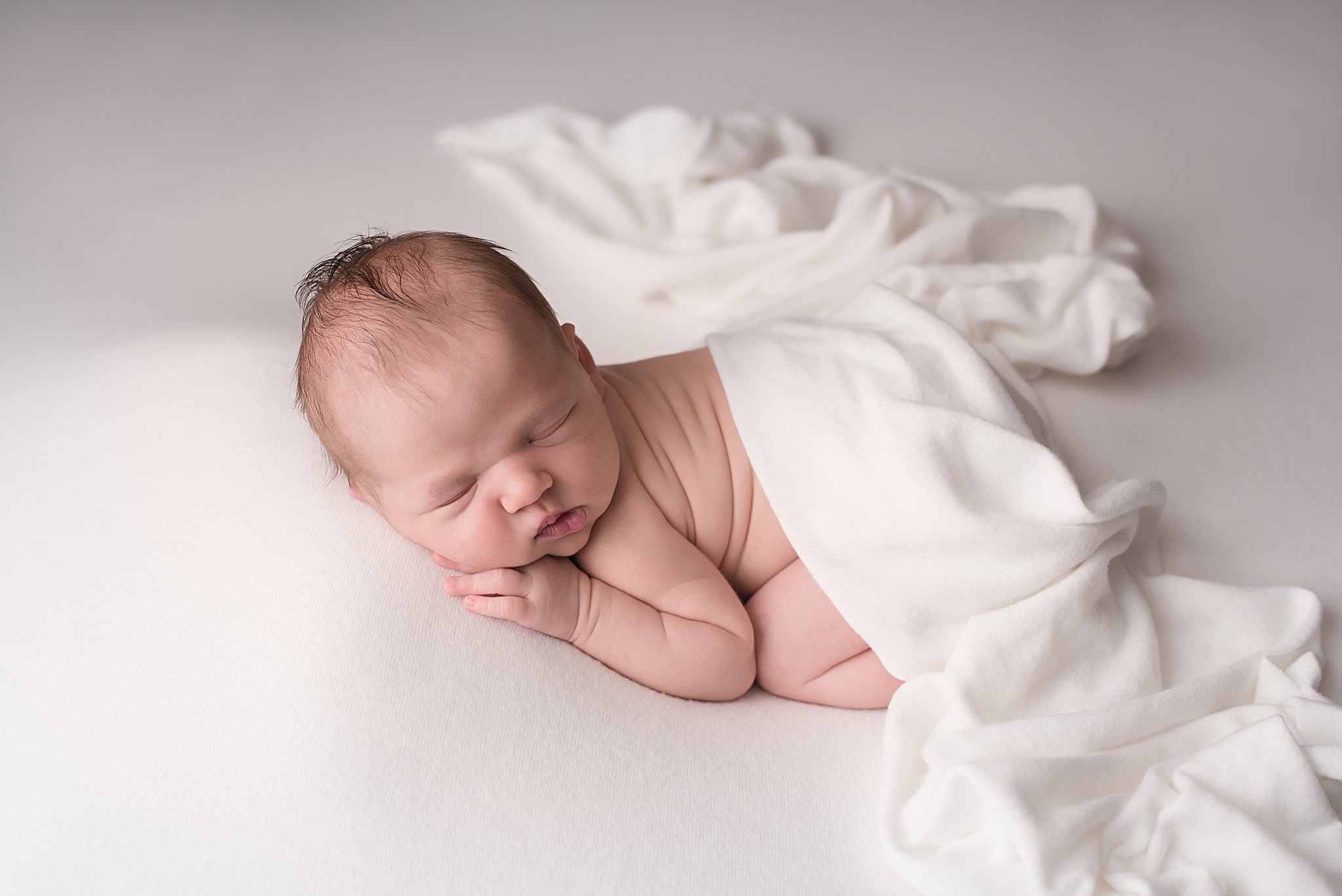 A newborn baby sleeps on a white bed with a white blanket draped over