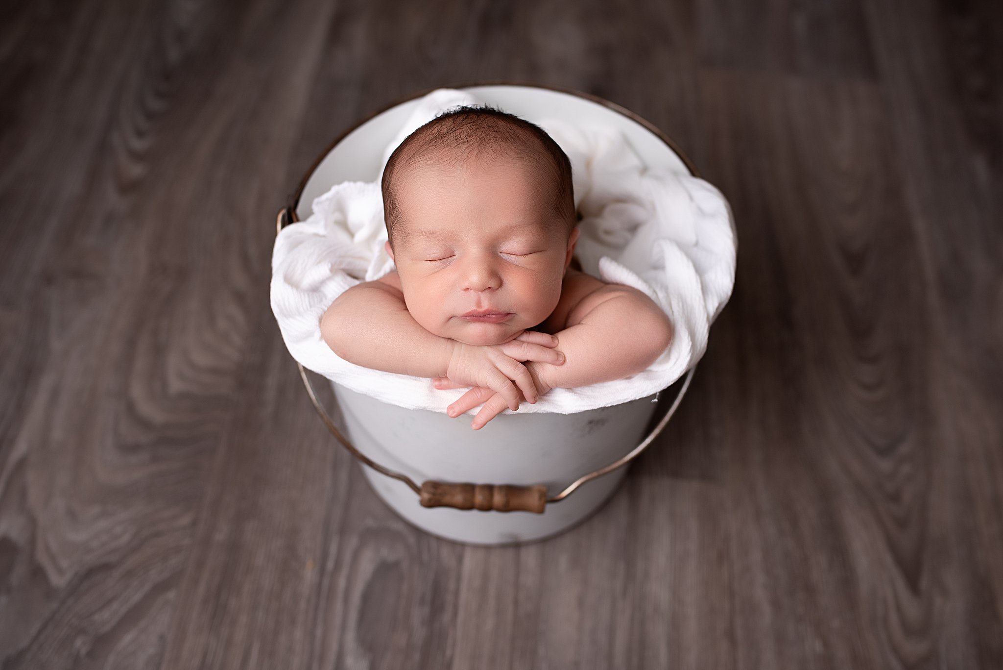 A newborn baby sleeps inside a metal bucket while resting its heads on its hands on the edge of the bucket baby's away san diego