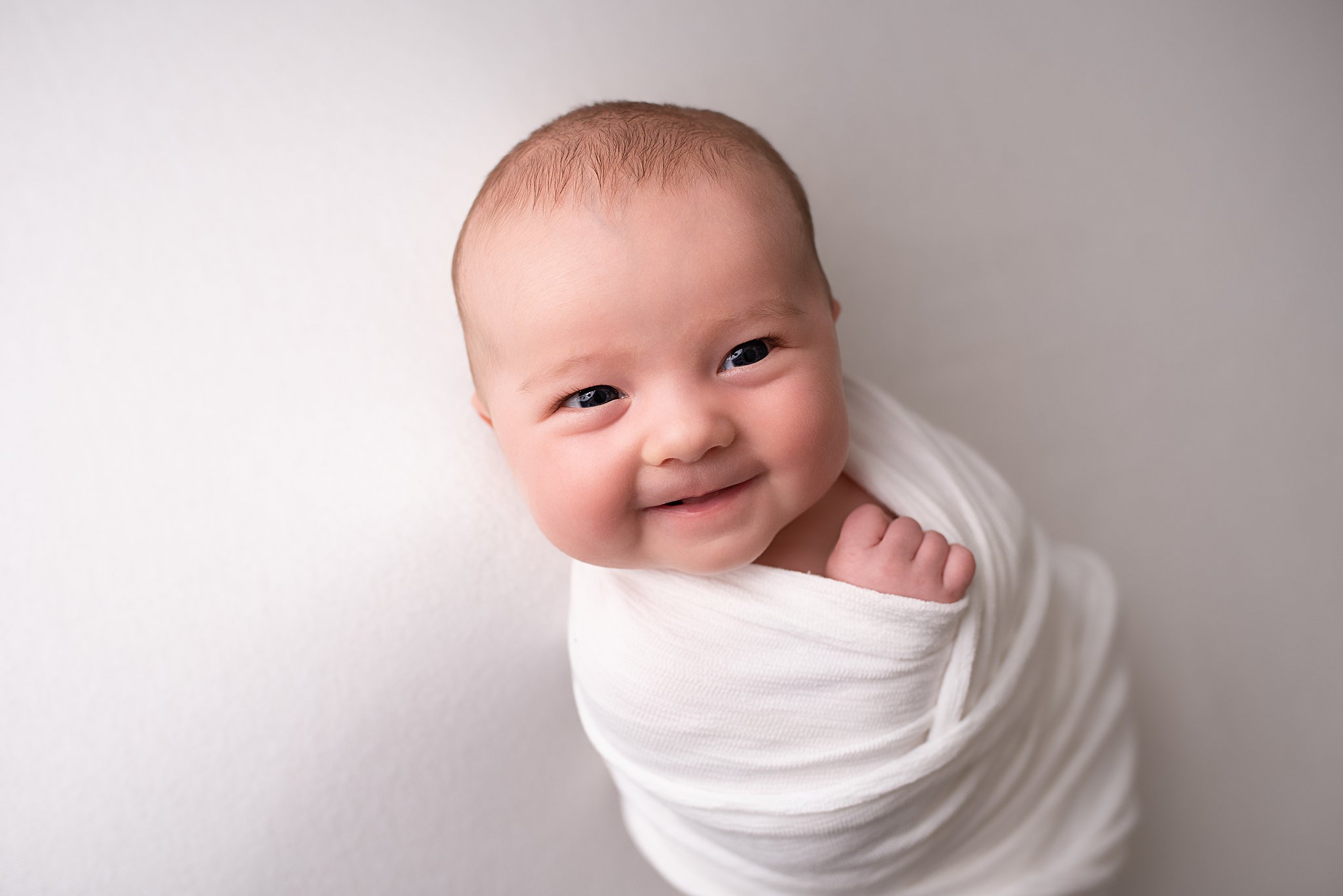 A newborn baby smiles while laying in a white swaddle