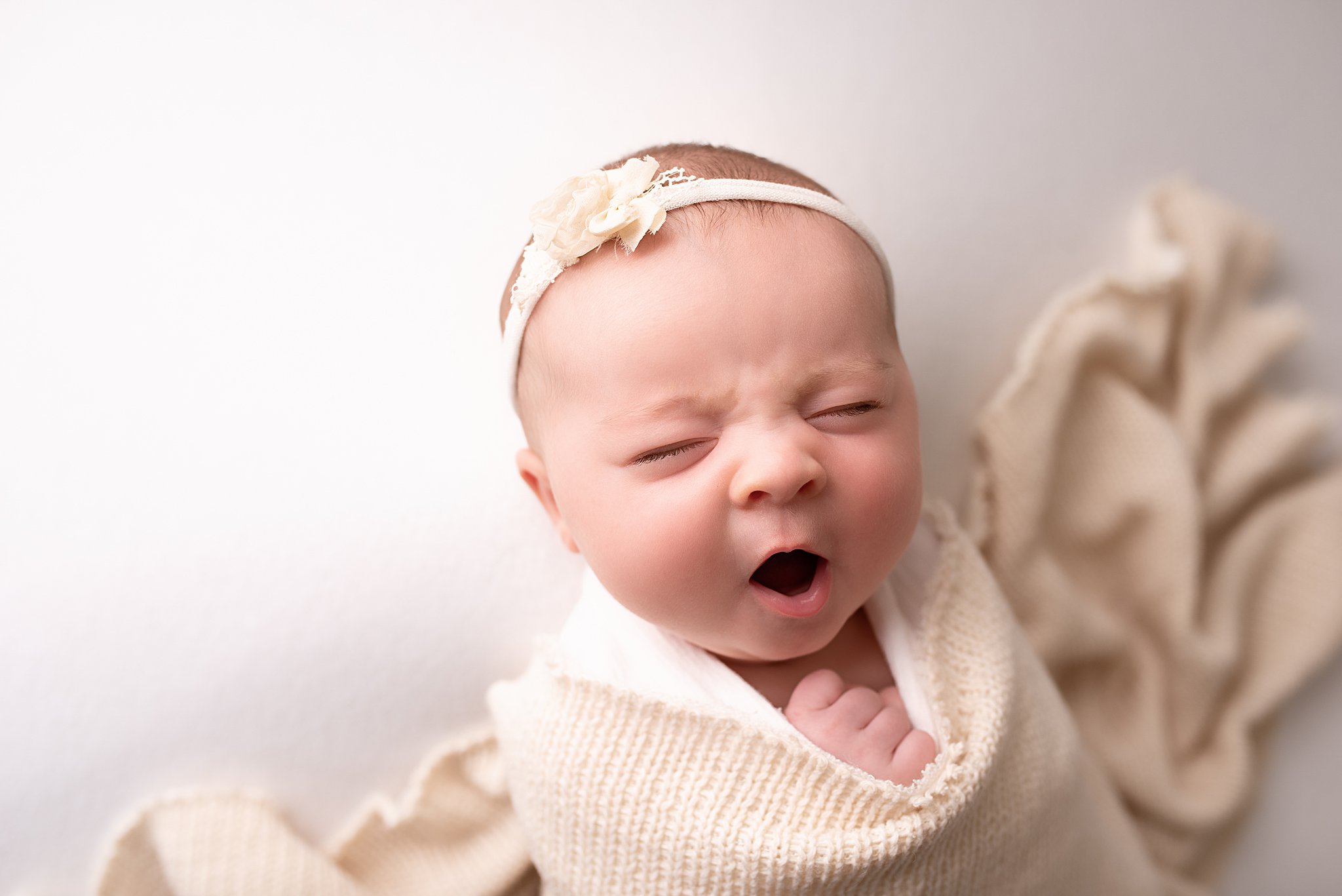 A newborn baby yawns while wrapped in a beige blanket luludew