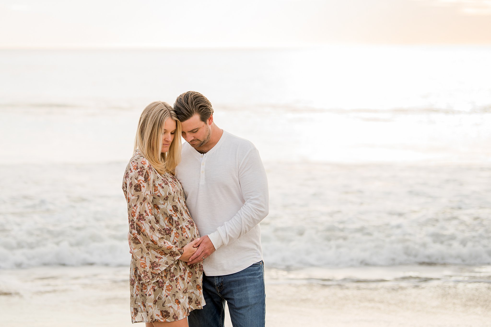A mother to be leans into her husband as they place their hands on her bump at sunset on the beach