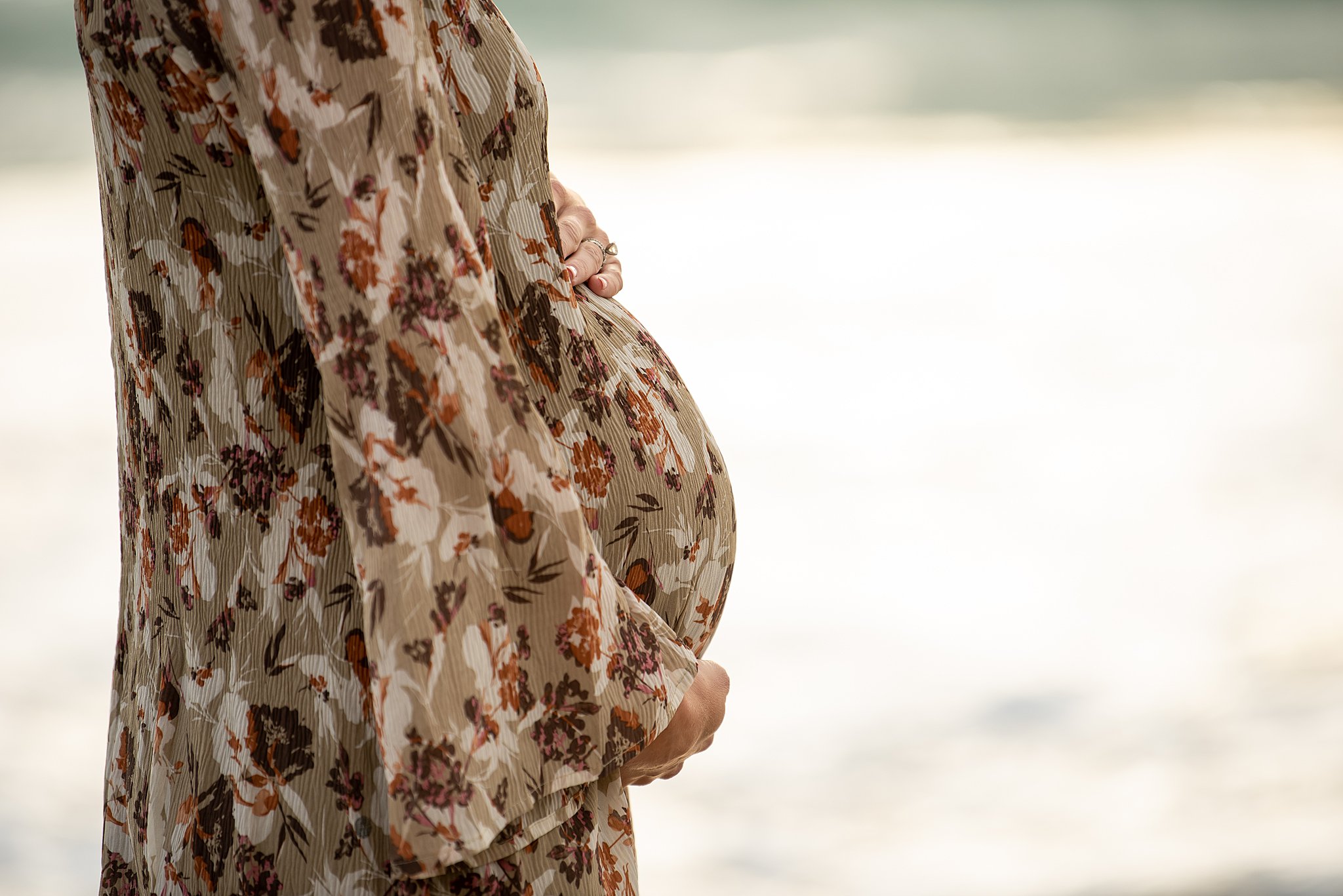 Details of a mother to be holding her bump in a floral maternity gown on a beach night and day San diego