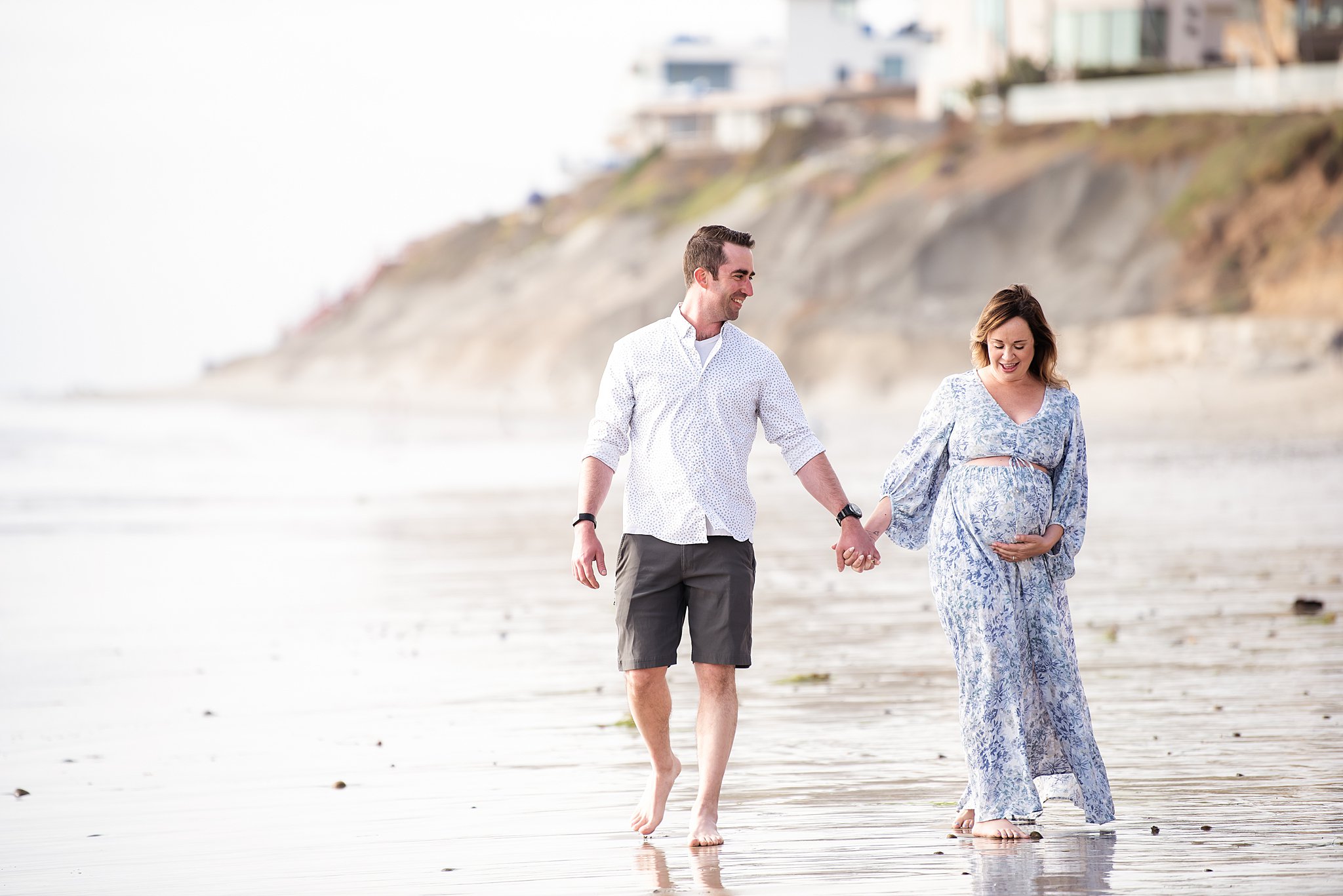 A mom to be holds her bump and her husband's hand as they walk down a beach together