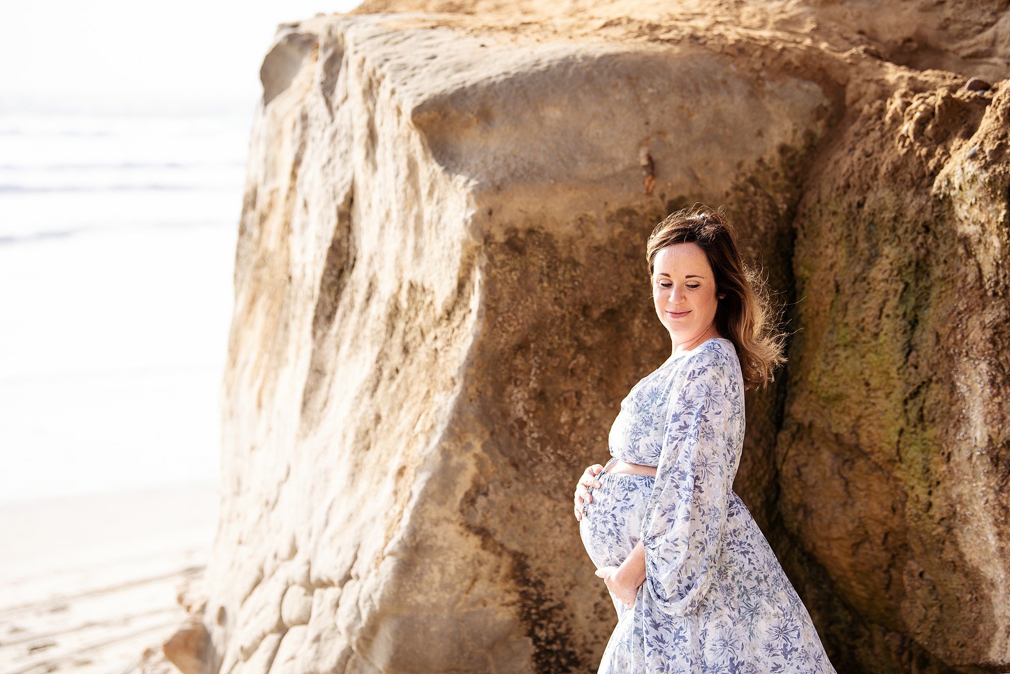 A mother to be in a blue floral dress places her hands on her bump on a rocky beach prenatal chiropractor san diego