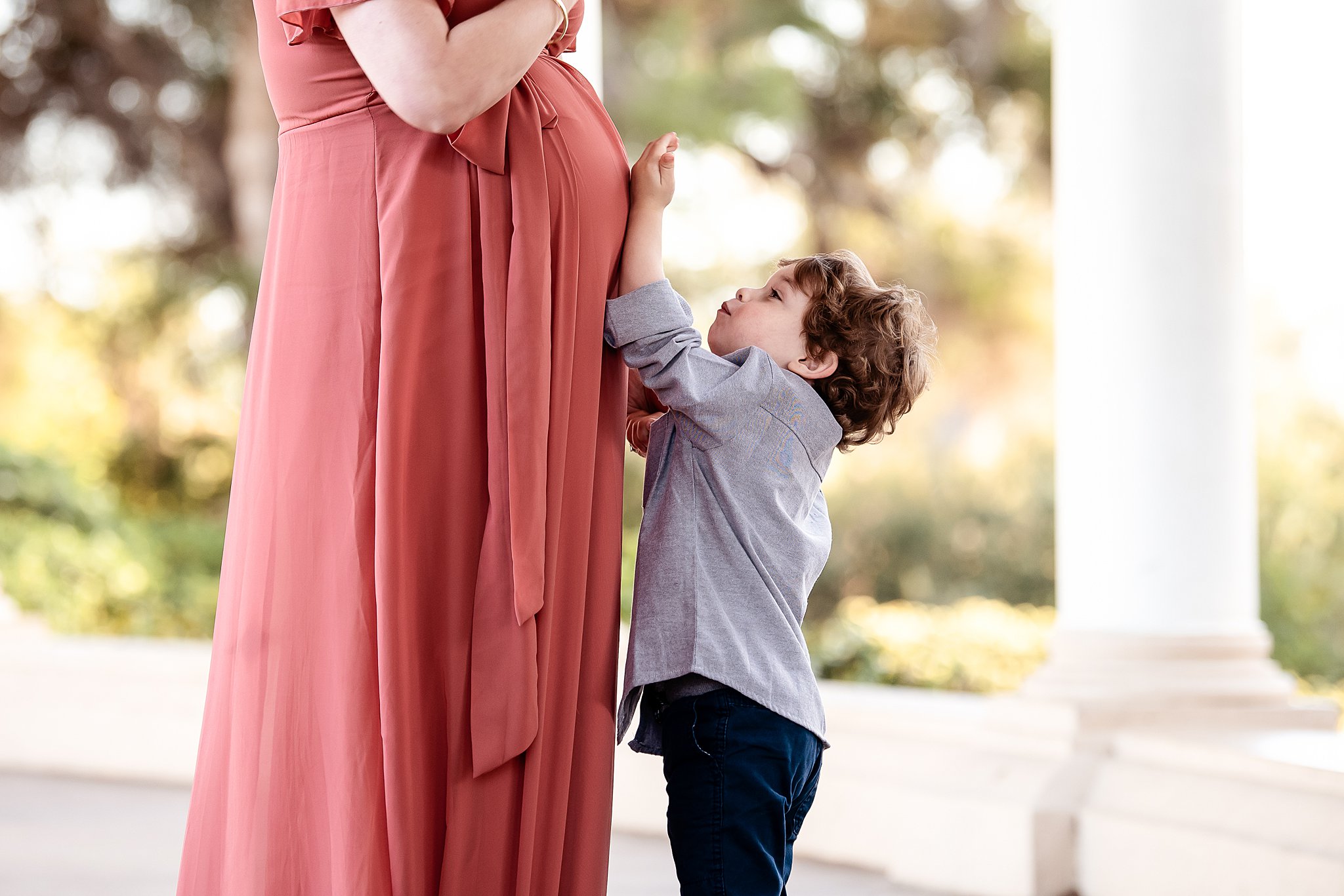 A young boy reaches up to his mom's pregnant belly in a park san diego baby shower venues
