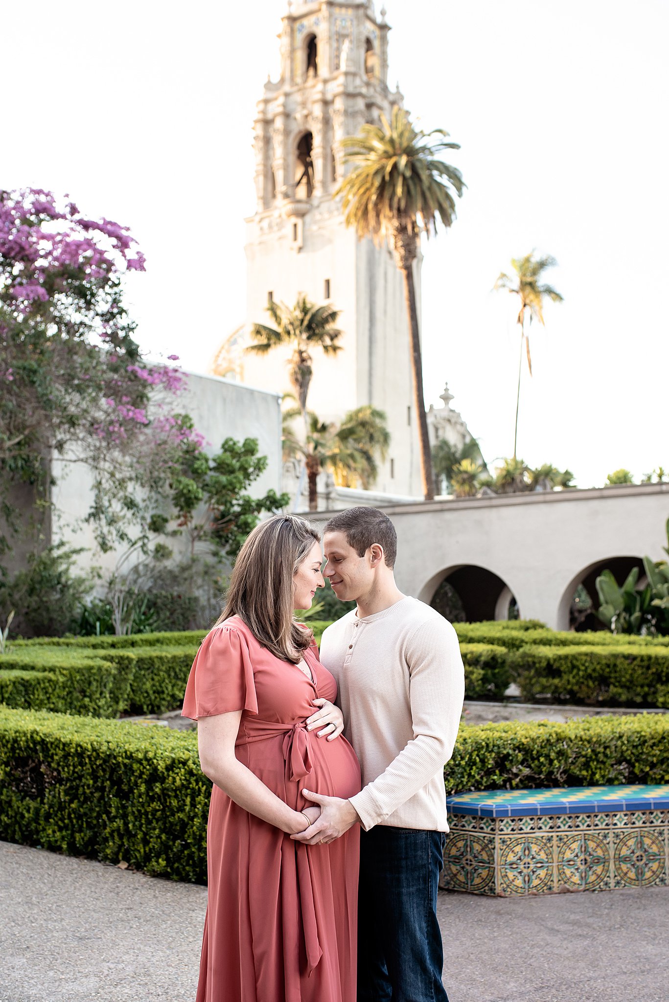 A mom to be in a pink maternity dress stands in a garden resting her forehead with her partners' san diego home birth