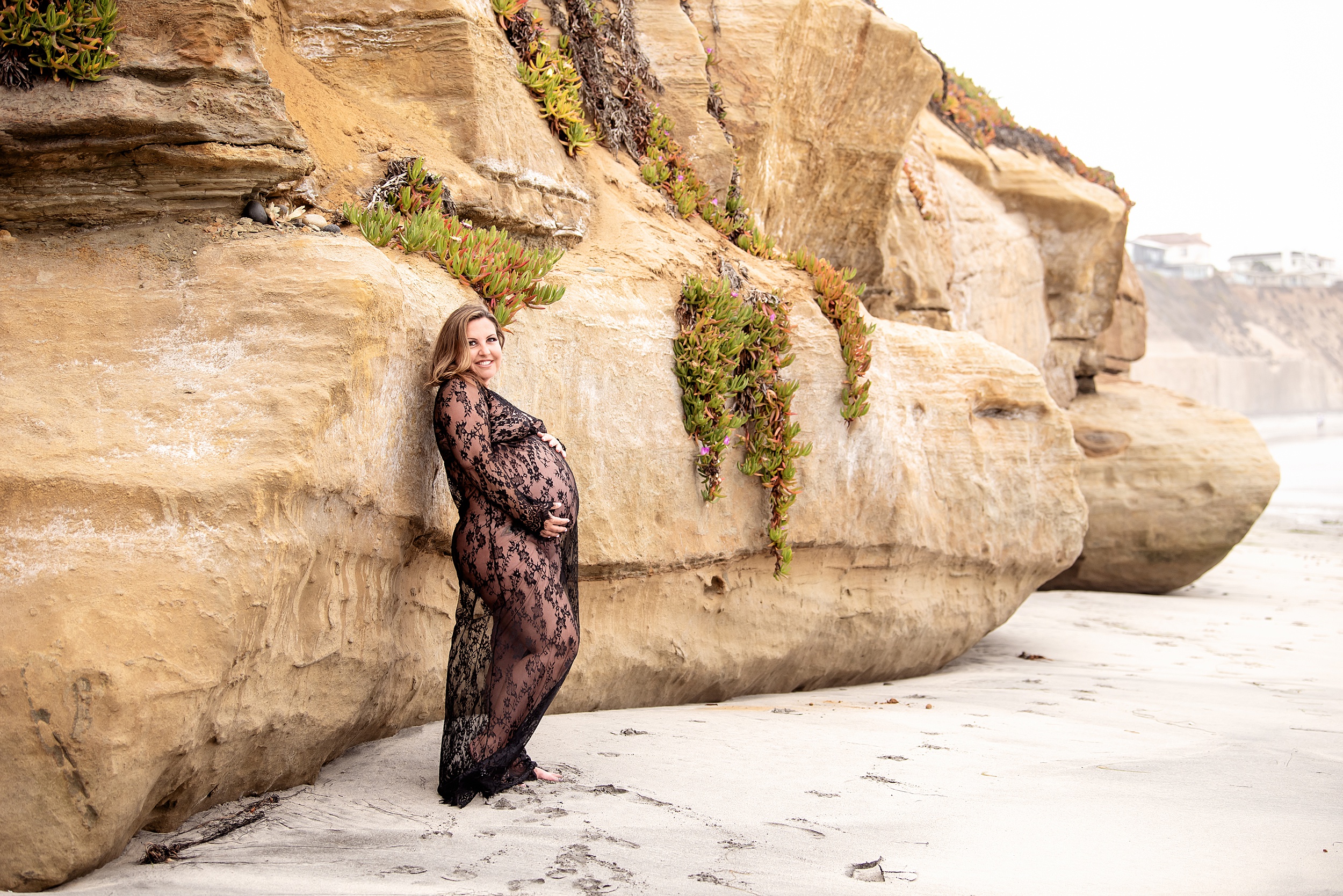 A mom to be in a lace maternity gown leans against a large rock on a beach Prenatal Pilates in San Diego