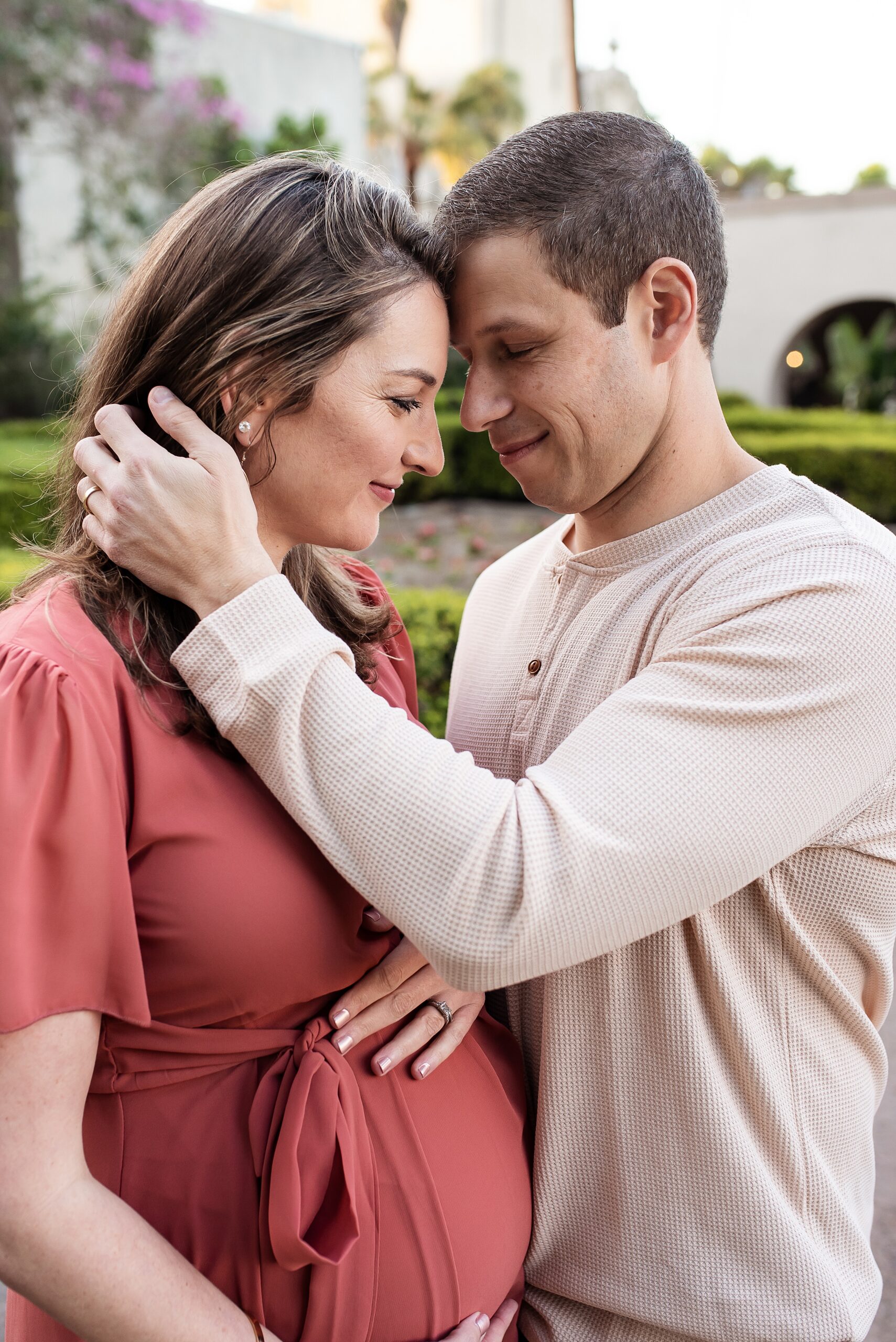 A mom to be holds her bump while pressing foreheads together while standing in a garden