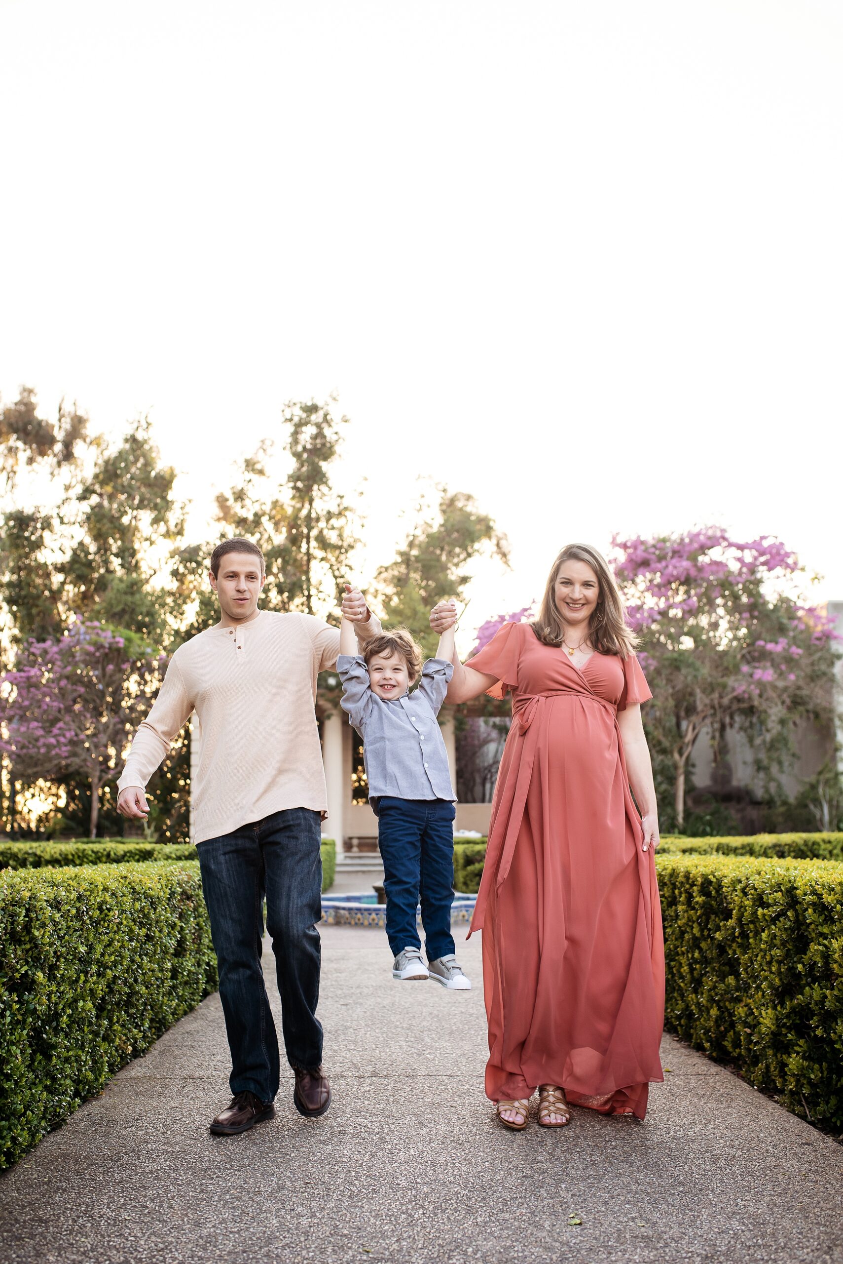 A pregnant mother and dad hold their toddler son's hands and lift him while walking through a garden path san diego 3d ultrasound