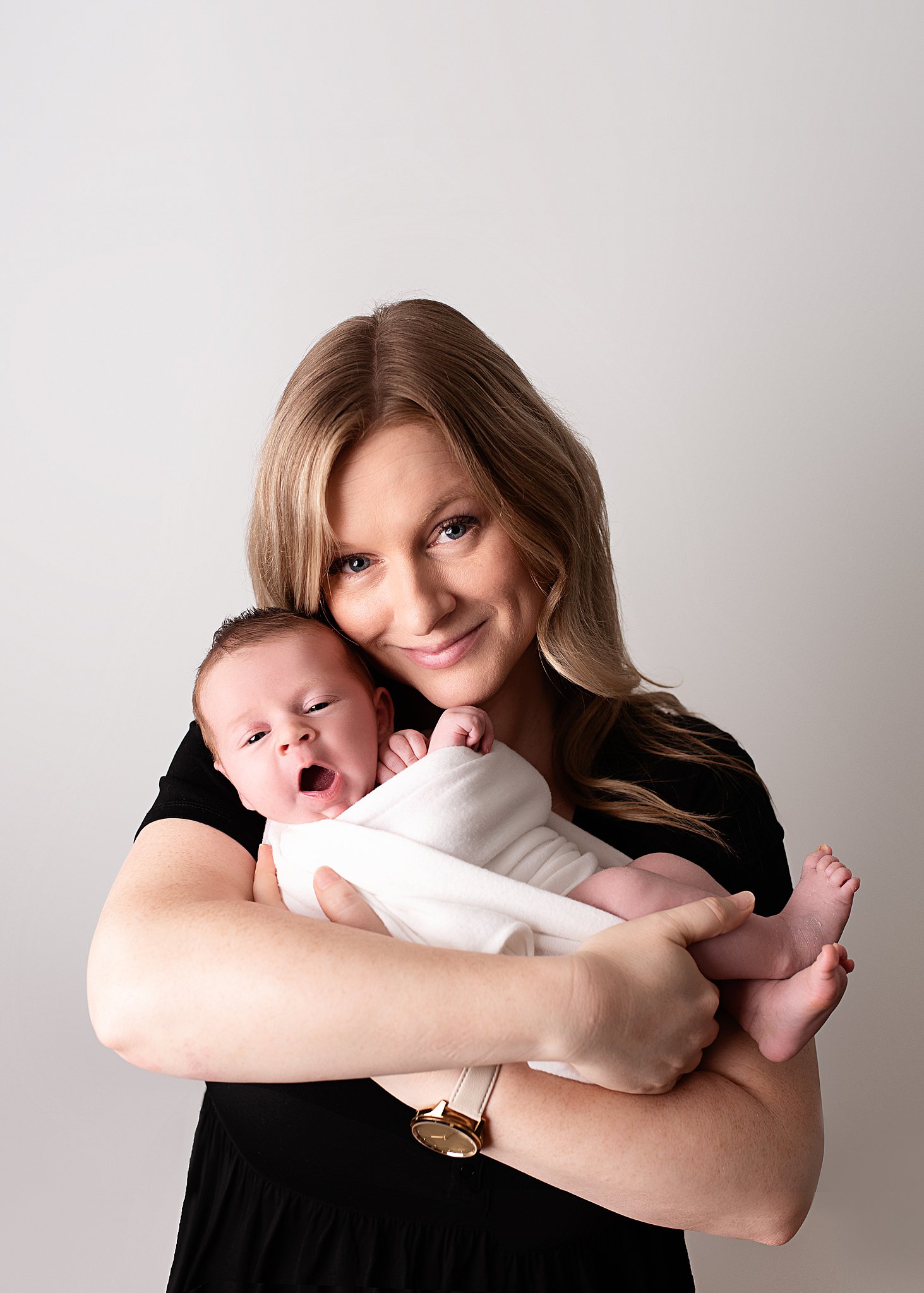 A new mom cradles her yawning newborn baby in her arms while standing in a studio