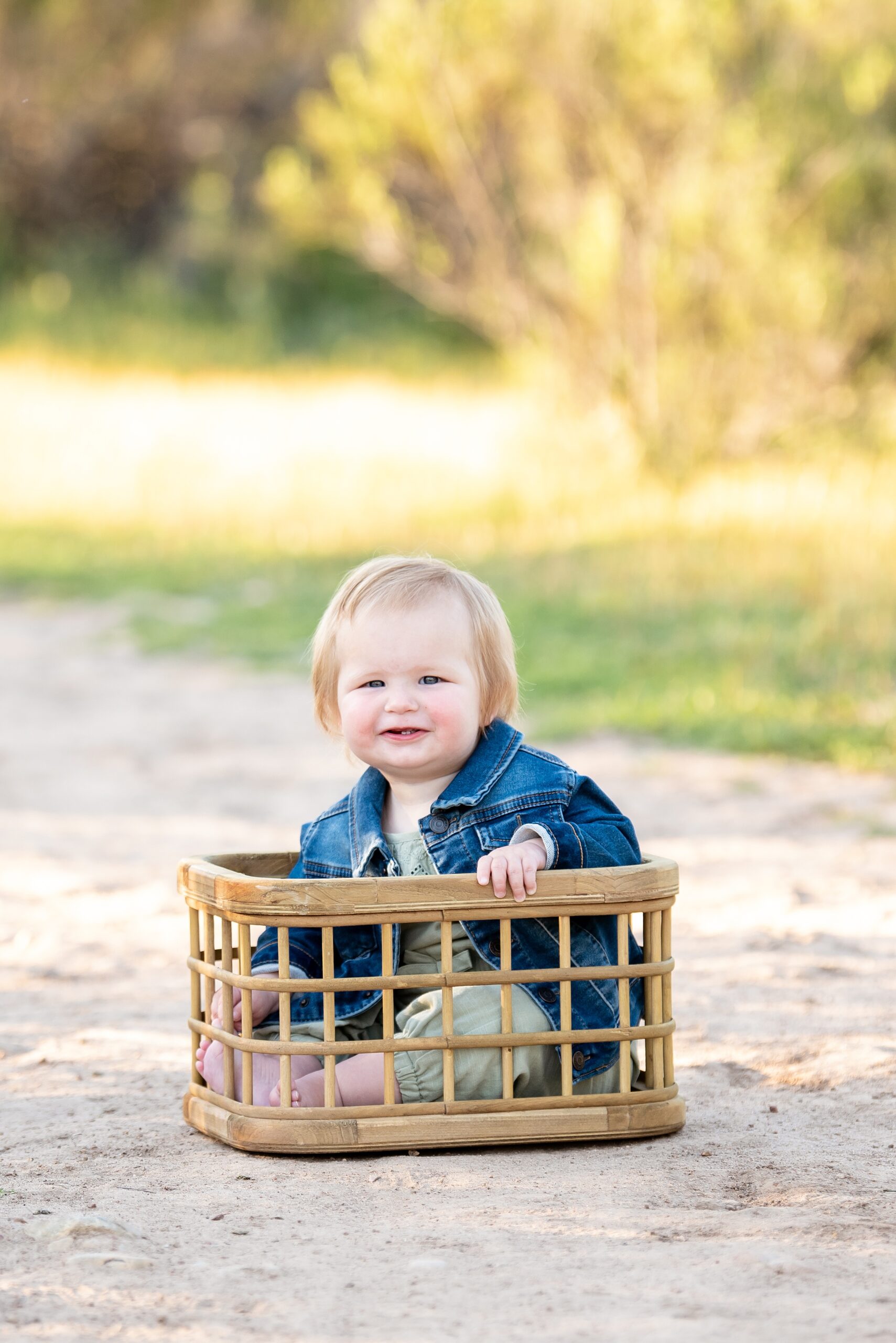 A young toddler smiles while sitting in a wooden basket in a park