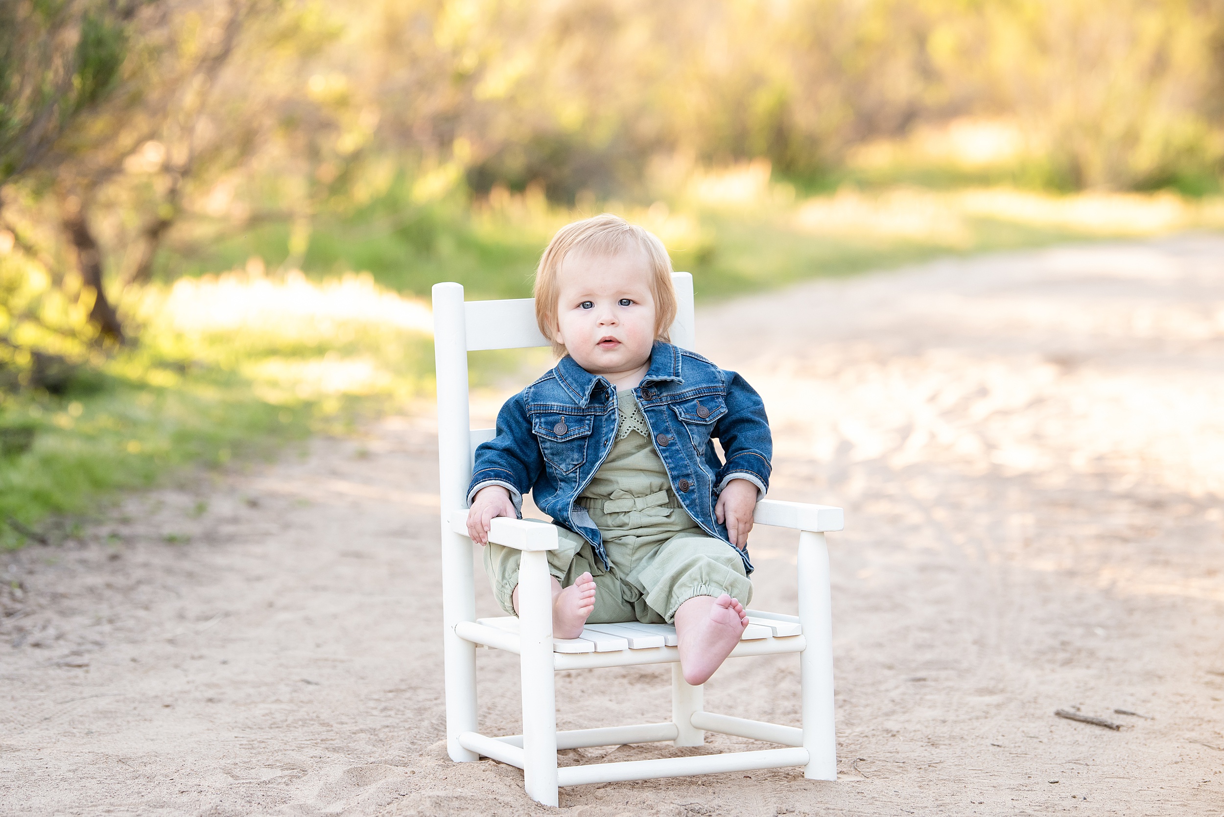 A young toddler in a denim jacket sits in a white chair in a sandy path at sunset after meeting a san diego au pair