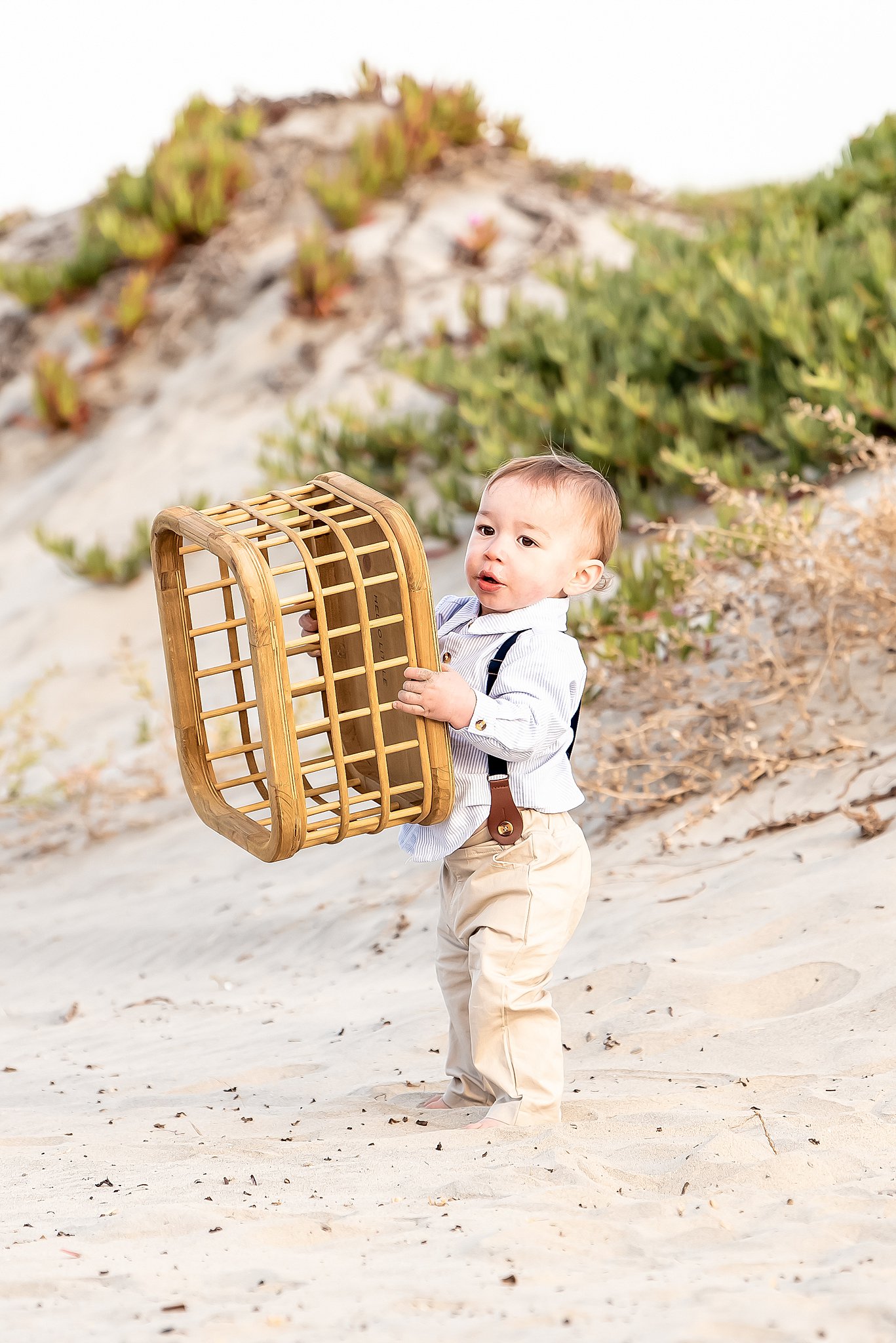 A toddler boy lifts a wicker basket while standing on a beach in khakis and suspenders after attending baby music class san diego