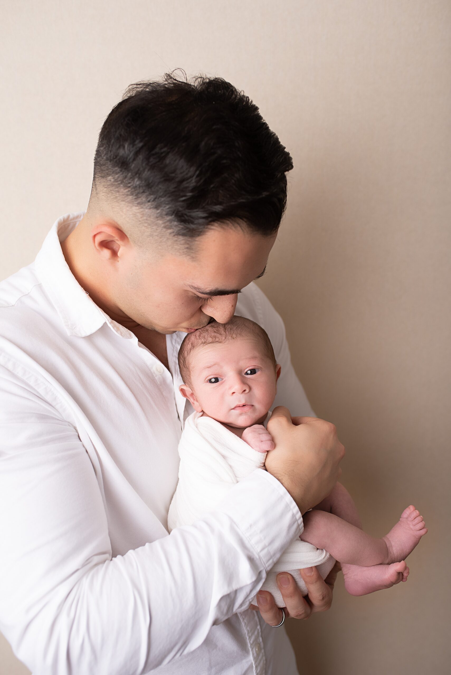 A father in a white shirt kisses the head of his newborn baby sitting in his hands against his chest after mom got san diego placenta encapsulation