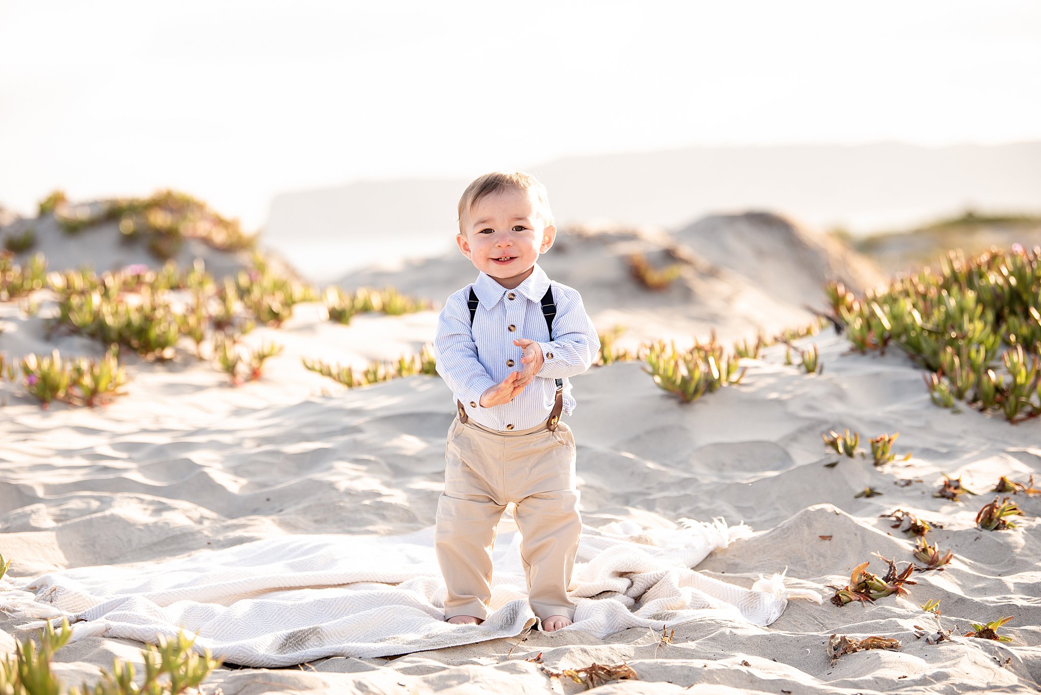 A toddler boy walks on a beach in khakis and suspenders at sunset before visiting san diego preschools