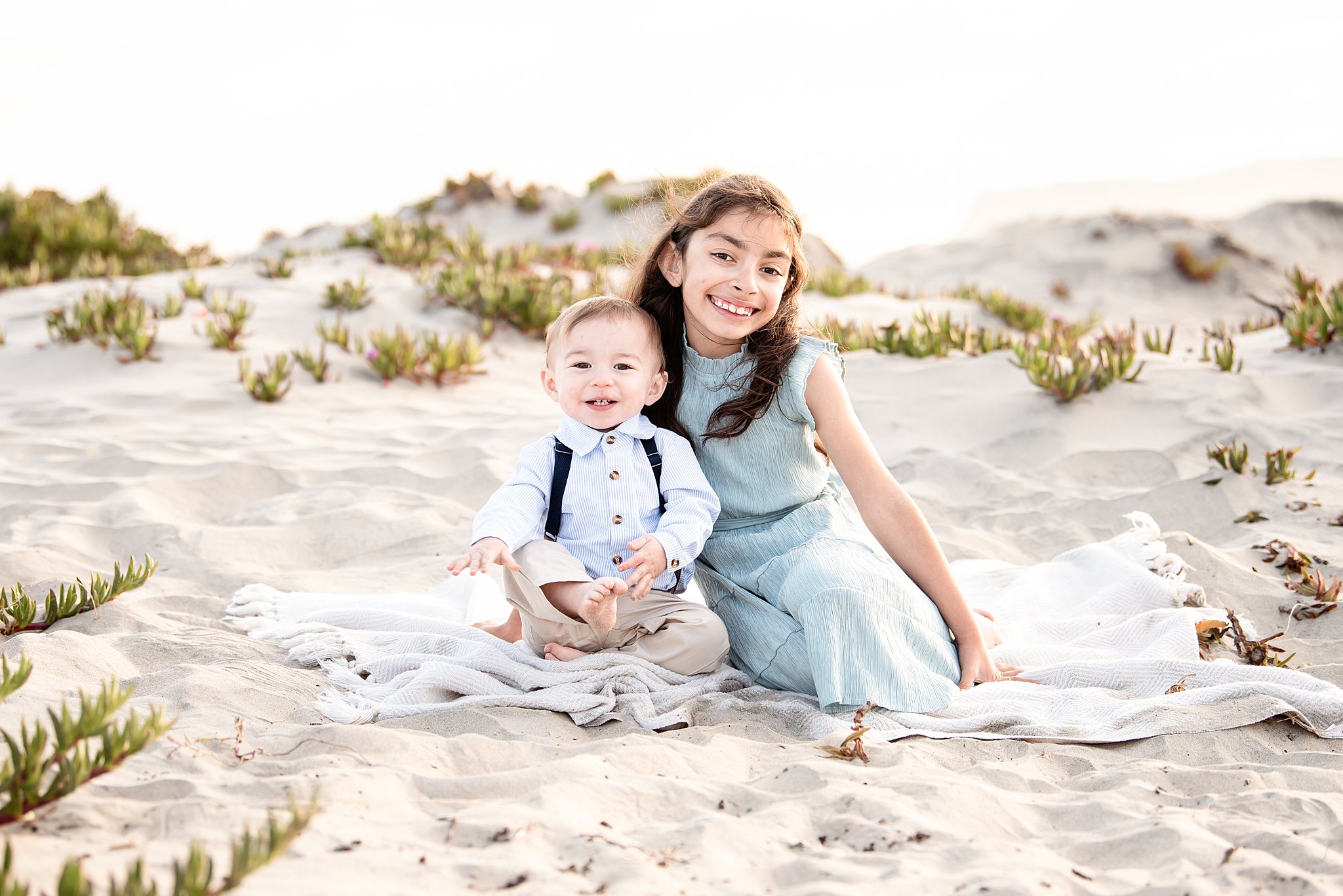 A young girl in a blue dress sits on a beach on a blanket smiling with her toddler little brother after attanding san diego preschools